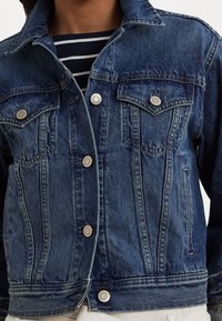 Denim jacket in dark blue with a classic collar, two chest pockets, silver buttons, and a slightly textured fabric. Contrast stitching detail.