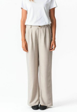 Vrouw draagt beige wijde broek, wit T-shirt met korte mouwen, zwarte textuurschoenen, staat met armen ontspannen langs het lichaam.