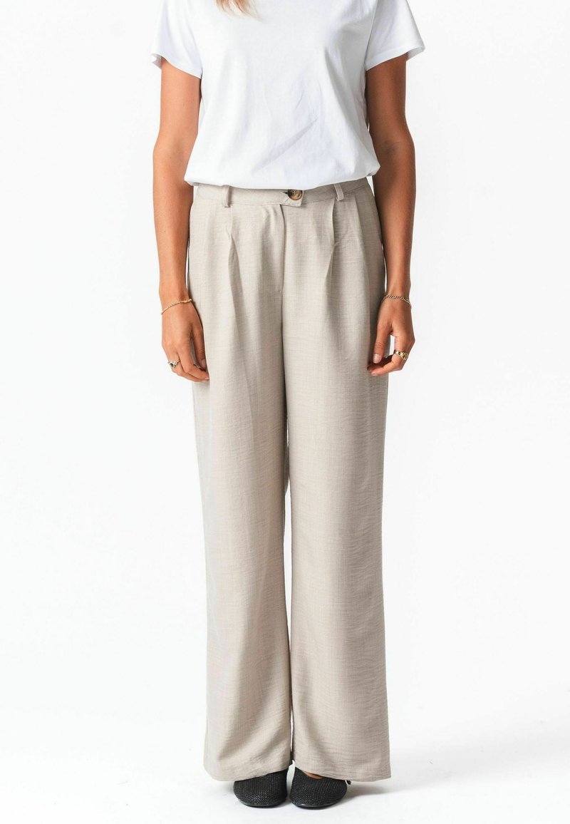 Femme portant un pantalon large beige, un T-shirt blanc à manches courtes, des chaussures noires texturées, debout les bras détendus le long du corps.