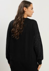 Mujer con largo cabello ondulado castaño lleva un suéter negro con pequeños puntos blancos en las mangas, vista desde la parte posterior y lateral.