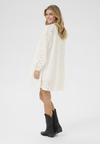 Vit klänning i ögonkänsla med långa puffärmar, rynkad fåll och utskurna blommönster; parat med svarta cowboystövlar.