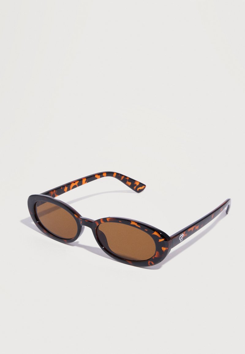 Οβάλ γυαλιά ηλίου με καφέ φακούς και σκελετό από χελώνια, που ακουμπούν σε λευκή επιφάνεια.