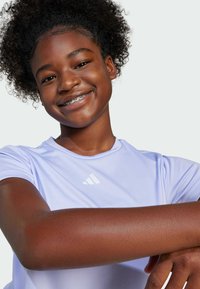 Camiseta deportiva de color púrpura claro con mangas cortas, que presenta un logo blanco de Adidas en el pecho. Textura suave y corte casual.