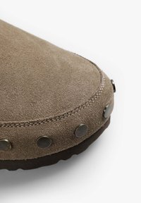 Zapato de ante marrón con punta redonda, que presenta tachuelas metálicas decorativas a lo largo del borde y una suela de goma oscura contrastante.