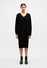 Rochie tricotată neagră cu decolteu în V, mâneci lungi și lungime până la jumătatea gambei. Asortată cu cizme negre până la glezne. Textură netedă și croi lejer.