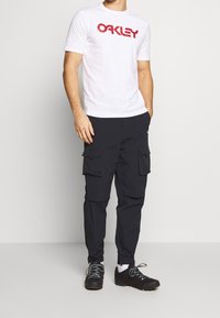 Bílá tričko s červeným logem "Oakley", spárované s černými cargo kalhotami se bočními kapsami. Černé boty s šedými tkaničkami dotvářejí outfit.