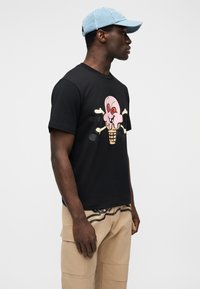 Μαύρο μπλουζάκι με γραφικό ενός ροζ χωνάκι παγωτού με σχέδιο κρανίου και σταυρωμένων οστών. Φοριέται με μπεζ παντελόνι καρό και ανοιχτό μπλε καπέλο.