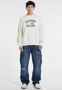 Felpa crema con la scritta "CALIFORNIA 81 GUESS JEANS" stampata in blu scuro. Indossata con jeans cargo blu larghi e strappati e sneakers bianche.