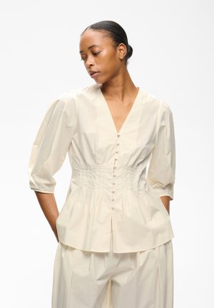 Femme portant un chemisier écru boutonné avec des manches bouffantes et une taille plissée, associé à un pantalon ample assorti, posant les mains dans les poches.