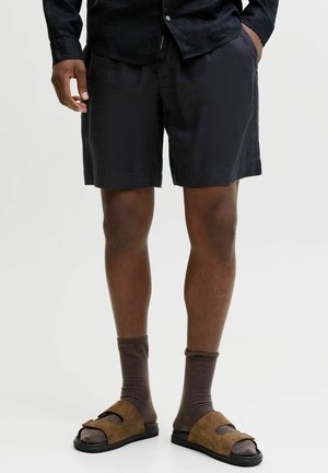 Moški v črnih kratkih hlačah, temni majici z dolgimi rokavi, rjavih nogavicah in rjavih sandalih z odprtimi prsti, ki stoji na enobarvni podlagi.