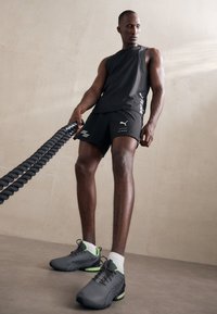 Svart ärmlös topp och shorts med vita detaljer. Skorna är grå med gröna detaljer. Personen håller i ett tjockt svart rep.