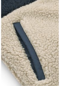 Tessuto shearling beige morbido con un pannello blu scuro liscio. Presenta una fessura per l'accesso alla tasca, evidenziando il contrasto di texture.
