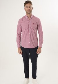 Camisa de cuadros rojos y blancos de manga larga con cuello abotonado, combinada con pantalones oscuros y zapatillas blancas. Presenta un pequeño logotipo en el pecho.