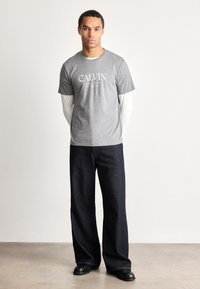 Młody mężczyzna stoi z rękami za plecami, ubrany w szary T-shirt z logo Calvin, pod którym ma białe długie rękawy, ciemne szerokie dżinsy i czarne buty.