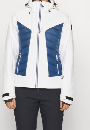 Person trägt eine weiß-blaue Jacke mit Reißverschluss und Kapuze sowie schwarze Hose, steht mit entspannten Händen an den Seiten.