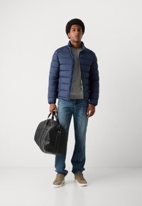 Veste rembourrée marine, t-shirt gris, jean bleu, baskets beiges. Sac de voyage en cuir noir avec double poignée. Textures lisses et lignes épurées.