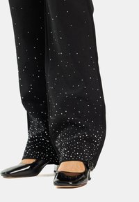 Pantalones negros con una textura suave, adornados con lentejuelas plateadas dispersas, combinados con zapatos negros de charol de punta cuadrada.