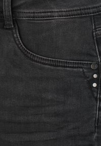 Gros plan d'une poche de jean en denim noir montrant les coutures, une boucle de ceinture et des rivets métalliques sur le tissu.
