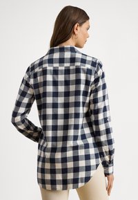 Károvaná košeľa v námorníckej modrej a bielej farbe, vyrobená z mäkkej látky. Má golier so zapínaním na gombíky, dlhé rukávy a zaoblený spodný lem.