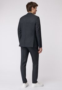 Dunkelgrauer Anzug, strukturierter Stoff, Slim-Fit, mit einem Schlitz hinten, kombiniert mit weißen Sneakers. Klare Linien und modernes Design.