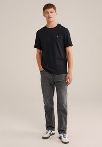 Jaunas vyras stovi su dešine ranka kišenėje, apsivilkęs juodą marškinėlį, pilkas džinsus ir baltus sportinius batus su juodomis juostelėmis prieš paprastą kreminį foną.