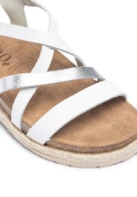 White leather sandal with metallic silver cross straps, a tan suede footbed, and a textured natural fiber sole. Smooth finish accents.