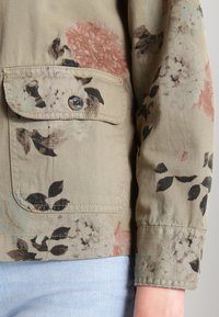 Blommönstrad jacka i khakifärgad tyg, med en ficka med knapp. Designen inkluderar dämpade rosa och svarta blommor samt en texturerad yta.
