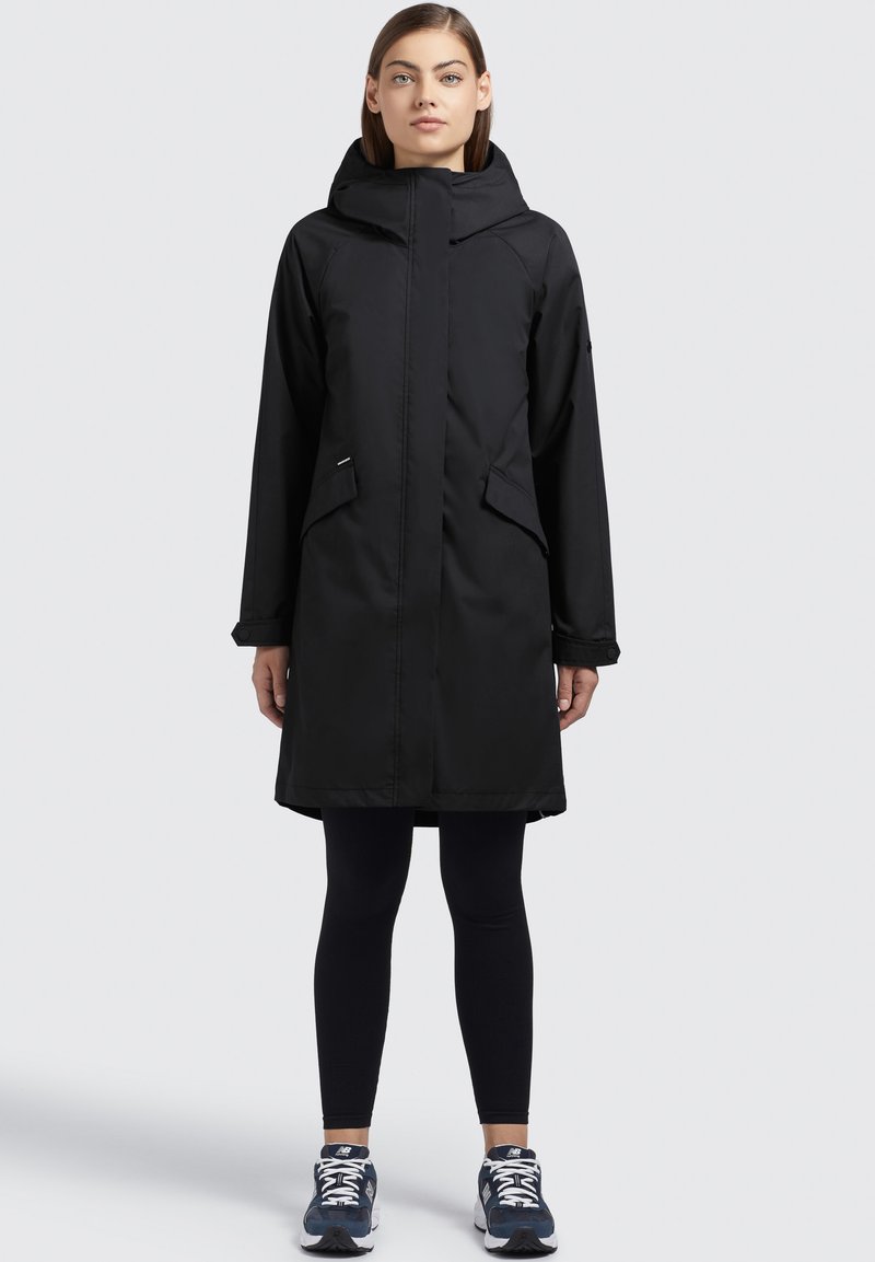 Veste imperméable noire de longueur genou avec capuche, deux poches avant, fermeture éclair et manches longues, associée à des leggings noirs et des baskets.