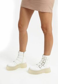 White ankle boots with a rounded toe, featuring three adjustable straps and silver buckles. Thick translucent sole adds height.