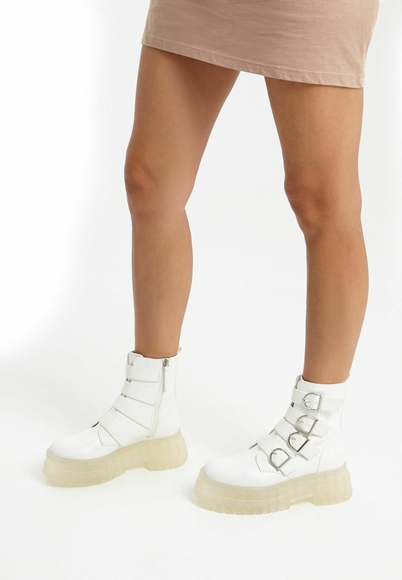 White ankle boots with a rounded toe, featuring three adjustable straps and silver buckles. Thick translucent sole adds height.