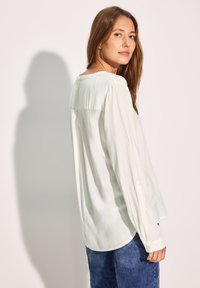 Blusa bianca a maniche lunghe con scollo rotondo e polsini con bottoni, abbinata a pantaloni in denim blu. Tessuto soffice e fluido.