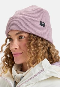 Roze geribbelde gebreide beanie met een opgevouwen rand en een zwart label met een hartlogo, gedragen met een lichtgekleurde jas.