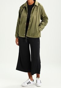 Osoba v olivově zelené košili na knoflíky, černých širokých zkrácených kalhotách, černém crop topu a bílých teniskách Adidas s černými pruhy.