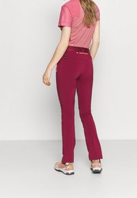 Pantalon de sport bordeaux en tissu extensible, doté d'une coupe ajustée, de jambes fuselées et de détails en zip orange contrastant. Associé à un haut rose.