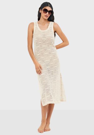 Mujer con vestido midi de crochet beige sin mangas, con abertura lateral y grandes gafas de sol redondas, de pie descalza sobre fondo blanco.