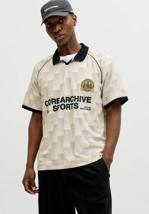 Homme portant un polo beige à manches courtes avec col noir et inscription "COREARCHIVE SPORTS", casquette noire et pantalon noir, debout devant un fond uni.