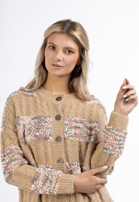 Tan stickad cardigan med flerfärgade texturerade lappar i rosa och blått, med fyra bruna knappar och ribbade ärmslut.
