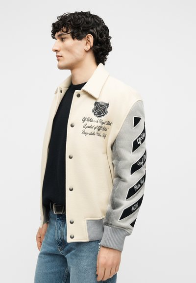 Νέος άνδρας με μια κρεμ και γκρι varsity ζακέτα με μαύρα γράμματα και διαγώνιες ρίγες στο μανίκι, φορώντας μπλε τζιν και μαύρο πουκάμισο.