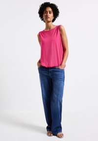 Top senza maniche rosa con dettagli arruffati sulle spalle, abbinato a jeans larghi blu. Il modello indossa sandali argentati e sta in piedi con le mani nelle tasche.