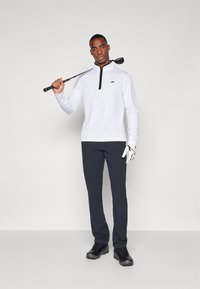 Pull-over de golf zippé blanc avec des accents noirs, associé à un pantalon gris foncé et des chaussures de golf noires, tenant un club de golf dans une main.