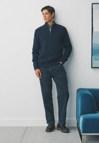 Hombre con suéter azul marino de media cremallera, camisa con estampado y jeans oscuros de pie, con una mano en el bolsillo, junto a un sillón azul y una obra de arte abstracto en la pared.