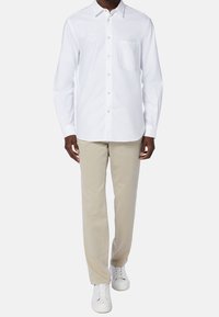 En person klädd i en vit skjorta med knappar, beige byxor och vita sneakers står mot en enkel bakgrund, med händerna längs sidorna.