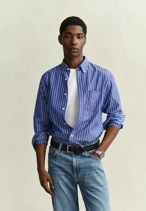 Jeune homme portant une chemise à rayures bleues et blanches boutonnée sur un t-shirt blanc, un jean bleu clair, une ceinture noire et une montre-bracelet argentée.