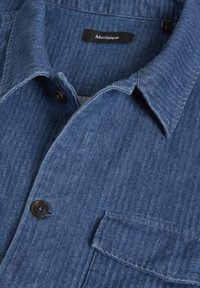 Camicia in denim blu con colletto a punta, caratterizzata da un motivo a spina di pesce, con bottoni marroni e una tasca sul petto. Etichetta del marchio visibile all'interno.