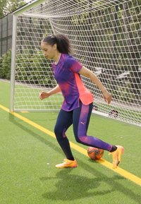 Roupa de desporto em violeta e rosa, estilo ajustado. Correndo num relvado verde com uma bola de futebol, usando chuteiras laranja e leggins pretos.