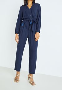 Navy blue jumpsuit with long sleeves, button front, a tie waist, and textured fabric. Accented with blue tassels on the belt.