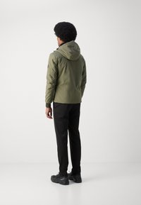 Doudoune vert olive avec capuche, équipée d'une fermeture éclair à l'avant et de poignets côtelés, associée à un pantalon noir et des chaussures noires texturées.