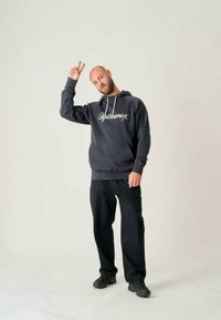 Tummanharmaa huppari, jossa on valkoiset vetonauhat ja logo, yhdistettynä mustiin housuihin ja kenkiin. Aihe viittaa kädellään rauhanmerkillä.