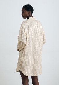 Beige gemustertes Oversized-Hemdkleid mit Kragen, das ein geometrisches Printmuster und einen abgerundeten Saum aufweist. Getragen mit dunklen Strumpfhosen.