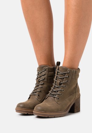 Brune læderstøvler med snøre ved anklerne og blokhæl båret på bare ben mod en hvid baggrund.